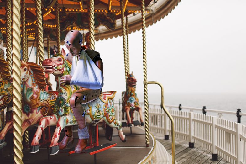 Crash test dummy with arm in a sling sitting on a merry-go-round. To illustrate mid-career academic scientists being weary of the precarity of their jobs and the constant merry-go-round of publications and job applications.