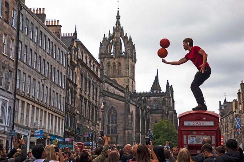 Street performer at the Edinburgh Fringe balancing basketballs on his finger while atop a former telephone kiosk bearing the word “cash”. To illustrate the precarious nature of Scotland’s university funding system.