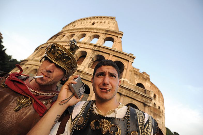 Two street entertainers dressed as ancient Romans listen to football in front of Rome’s Colosseum gladiators