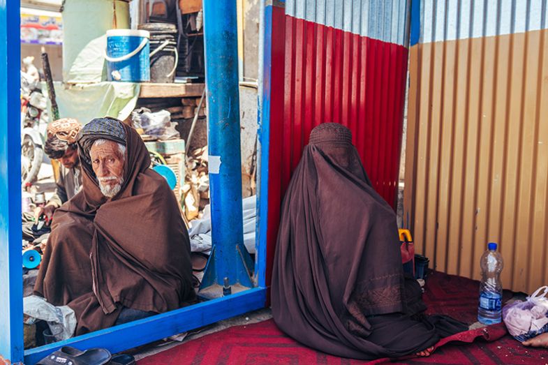 An elderly man and a woman in a burqa, sitting back to back on a city street, Kandahar, Afghanistan, April 2025. 