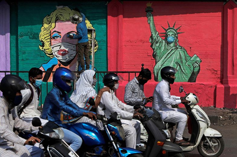 People on mopeds drive past an artist painting a mural of Marilyn Monroe, beside one of the Statue of Liberty wearing face masks in Mumbai, India, 2021. To illustrate Asian students being put off studying in the US, especially after Covid.