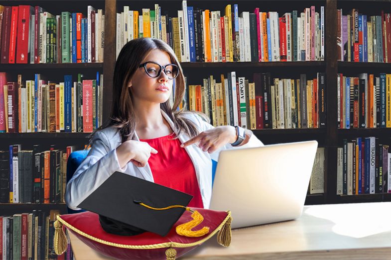 Student pointing at herself in a library, with a laptop and a mortar board on a velvet cushion on her desk. To illustrate online learning and student-centred teaching.