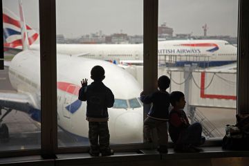 Children looking at British Airways planes out of an airport window. To illustrate the UK banning dependants. 
