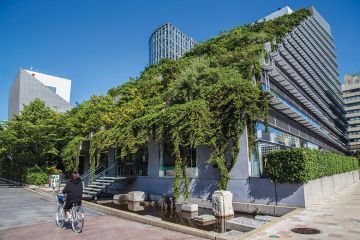 woman cycles past modern building in Japanese city with green roof of plants