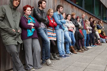 Crowd of grumpy looking students leaning against a wall, to illustrate a rise in student complaints.