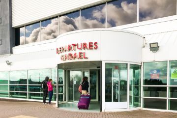 Cardiff, Wales - June 2019:: Person pulling a suitcase entering the terminal building at Cardiff Wales Airport. Blue sky and clouds are reflected in the windows. The airport is publicly owned by the Welsh Government.