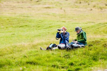 Two senior adults, a man and a woman, sit on the ground in a green meadow in Skye, Scotland