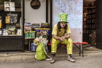 A man in a clown costume sitting and smoking cigarettes in front of an antique bookstore.