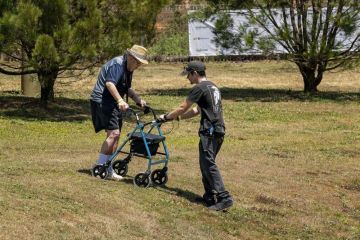 A young man assists and elderly male with walking frame navigate himself down a hilly slope at a country fai