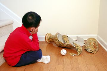 A young boy contemplates a smashed vase, with a ball sitting next to it A young boy contemplates a smashed vase, with a ball sitting next to it