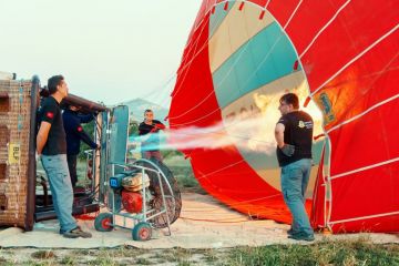 Mens inflating of hot air balloon with large fans, basket lies on the ground, preparation for the upcoming flight against distant hills, early morning clear sky