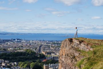 Standing on a rocky precipice, a person overlooks urban scenery and the ocean, under a bright sky