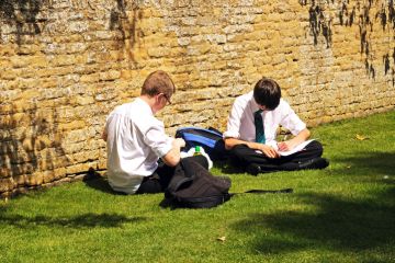 Two teenage schoolboys sitting on the grass by a Cotswold stone wall doing their schoolwork.