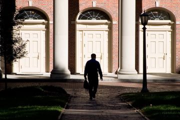 A man hidden in shadows walking towards entrance of building. To illustrate that staff accused of sexual harassment are free to leave their position and gain a new job at a different institution with no obligation to disclose any allegations.