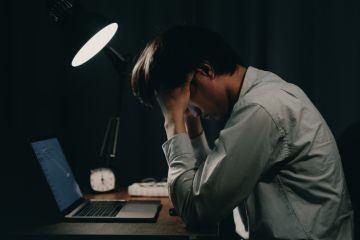 A man working on a laptop holds his head late at night, illustrating overwork
