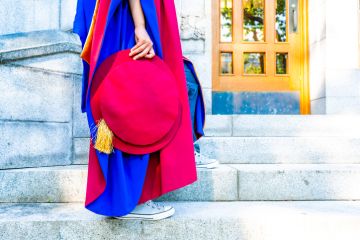 PhD (doctoral) graduate in regalia gown, holding Tudor bonnet cap, sitting on university steps, with sneaker canvas shoes showing. Red and blue grad gown, gold tassel showing.