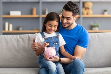 Portrait of smiling little girl putting coin in pink piggy bank, sitting on dad's lap on the couch at home