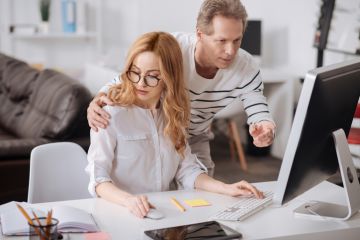 A man touches a woman's shoulder as she works on her computer, symbolising sexual harassment