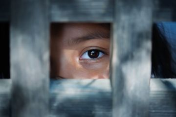 The blank stare of the eye of a child who is standing behind what appears to be a wooden frame illustrating modern slavery