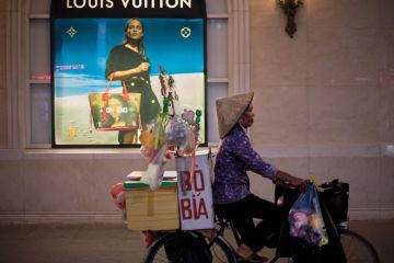 A bicycle vendor offering her merchandise in the streets of Hanoi pedals past a high end merchandise mall in the center of the city to illustrate Cost ‘barrier’ for Vietnam branch campuses under revised rules