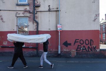 Two men carry a mattress over their heads past a terraced house, the walls of which bare graffiti reading "food bank" to illustrate Cost of living crisis bites for PhDs on ‘inadequate’ stipends