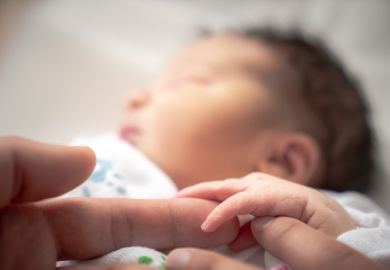 A newborn infant baby girl in a blanket swaddle wraps her tiny hand and fingers around her father and mother's fingers as she sleeps peacefully.