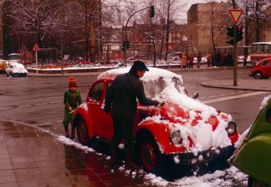 Berlin (West), Germany, 1977. Together, father and son remove snow from the family vehicle at a crossroads. Under the snow, a red VW Beetle hides.