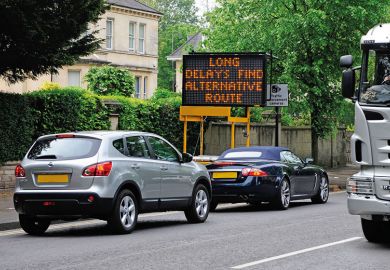 Cars in traffic jam with 'Long delays, find alternative route' sign