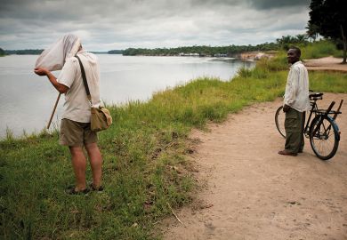 A man looks on in amusement as Ashley Kirk-Spring, a British scientist studying flies beside the Lomami River in DRC, has to put his head right into the net he has caught them in to examine them closely