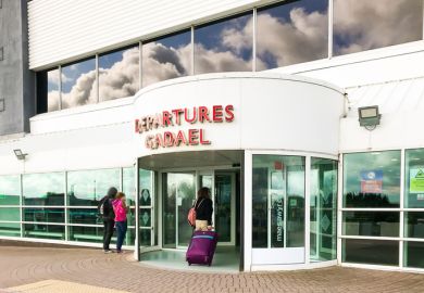 Cardiff, Wales - June 2019:: Person pulling a suitcase entering the terminal building at Cardiff Wales Airport. Blue sky and clouds are reflected in the windows. The airport is publicly owned by the Welsh Government.