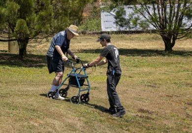 A young man assists and elderly male with walking frame navigate himself down a hilly slope at a country fai A young man assists and elderly male with walking frame navigate himself down a hilly slope at a country fai
