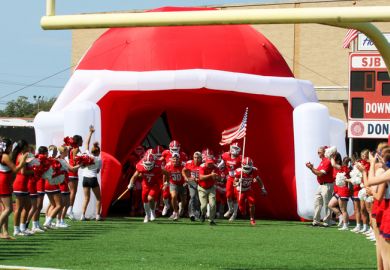 High school football team entering the field through a blow up football helmet tunnel with cheerleaders in a line cheering.