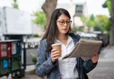 A young woman in the street holds a coffee cup in one hand and a newspaper in the other