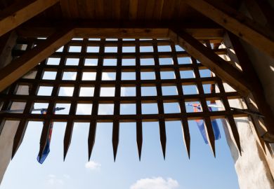 View of a medieval wooden portcullis gate with blue sky and flags in the background