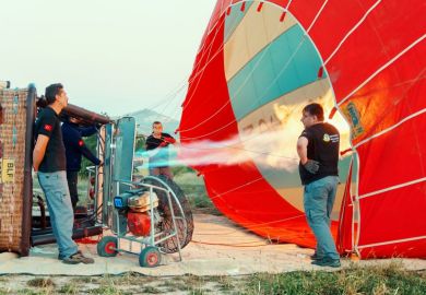 Mens inflating of hot air balloon with large fans, basket lies on the ground, preparation for the upcoming flight against distant hills, early morning clear sky