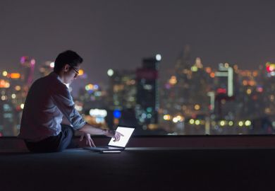 A man in a dark room overlooking a cityscape at night uses a laptop