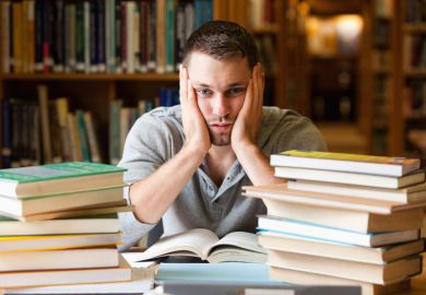 In a library, a student behind a pile of books clasps his face in his hands