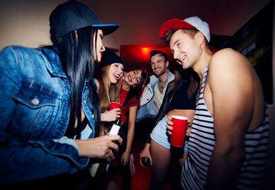 Low angle group of young stylish people standing in dark hall of nightclub together, talking, laughing and drinking beer as they wait to be admitted