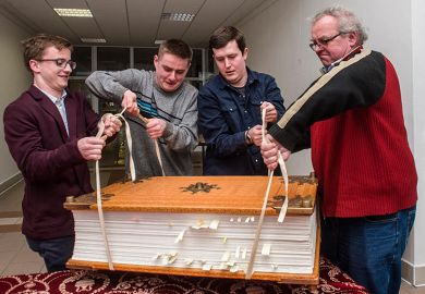 Men struggling to lift a giant book. To illustrate that three major research universities have opted out of new Elsevier deal amid concerns over the cost.