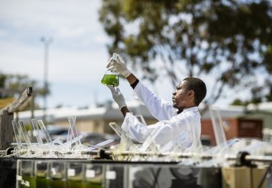 A young scientist working on research outdoors, holding up a glass beaker and inspecting the liquid inside, symbolising open science 