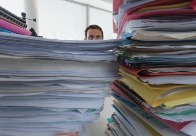 A man peers out from behind a pile of paperwork