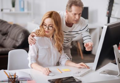 A man touches a woman's shoulder as she works on her computer, symbolising sexual harassment
