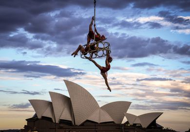 Trapeze performers hang from a cable in front of the Sydney Opera House