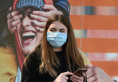 A woman wearing a face mask stands by an advertising placard showing someone having their eyes covered, April 21, 2020