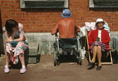 Ageing, elderly parents sunbathe with a teenage daughter as the father oddly faces a brick wall while sat in his wheelchair. Looking bored with the family holiday, the young lady of about 18 years of age, sits on a concrete block