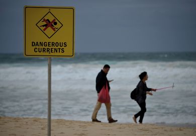 Visitors walk on Bondi beach next to a warning sign for dangerous sea currents on a stormy day in Sydney