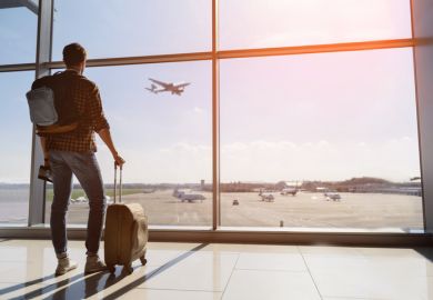 A traveller with a suitcase looks out from an airport terminal building at a plane taking off