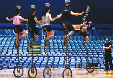 five people on unicycles balancing bowls on their heads with one toppling down as a metaphor for enrolments uncertainty in Australian Universities 