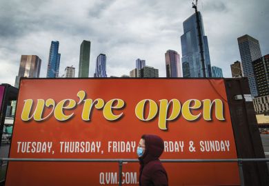 A woman wearing a face mask walks past a billboard reading 'We're open' in Melbourne, Australia to illustrate Australian universities head back to campus after shutdowns