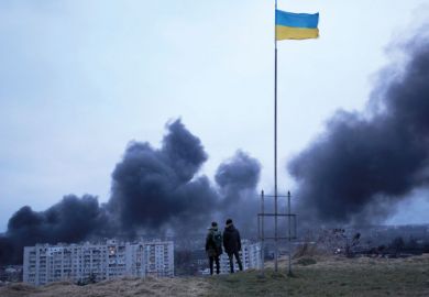 People standing near a Ukrainian national flag watch as dark smoke billows following an air strike in Lviv to illustrate Ukraine universities teach under fire